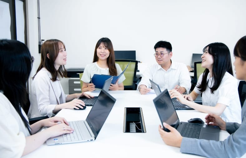 写真：オフィス内でスタッフ6名が話し合っている様子。和やかな雰囲気。